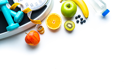 Overhead view of a composition of healthy fruits, weight scale, tape measure and dumbbells shot on white background. The composition is at the top of an horizontal frame making a border and leaving copy space.  High resolution 42Mp studio digital capture taken with SONY A7rII and Zeiss Batis 40mm F2.0 CF lens