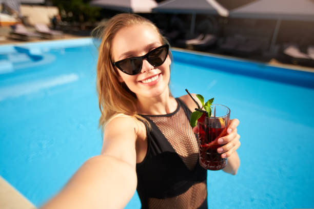 Travel blogger woman in bikini taking selfie photo with cocktail near swimming pool. Pov of lifestyle vlogger selfie from vacation on luxury resort. Female in swimwear drinks beverage on tropical spa. stock photo