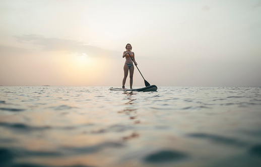 Woman silhouette paddling SUP board on sunset sea.