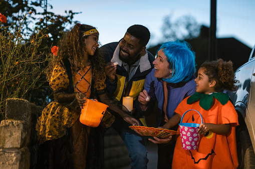 A family wearing fancy dress, out trick or treating in North East England during halloween. They are standing on a footpath, holding buckets and marshmallows while all looking at the eldest daughter excitedly.