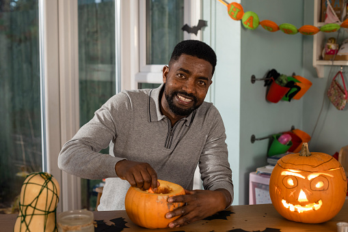 A man sitting at his kitchen table at home in North East England during halloween. He is carving a pumpkin while looking at the camera and smiling.