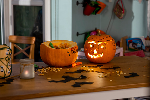 A dining table in a kitchen in North East England during halloween. There is a carved pumpkin and a partially decorated pumpkin on the table surrounded by pumpkin seeds and handmade paper bats.