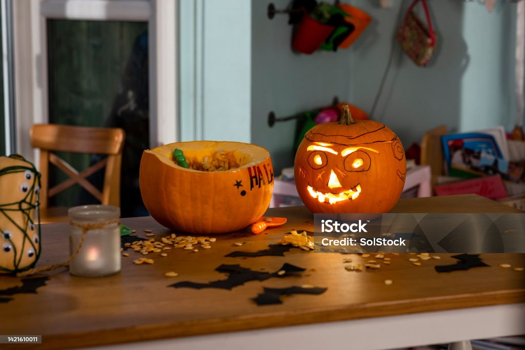 Scary Homemade Pumpkins A dining table in a kitchen in North East England during halloween. There is a carved pumpkin and a partially decorated pumpkin on the table surrounded by pumpkin seeds and handmade paper bats. Jack O' Lantern Stock Photo