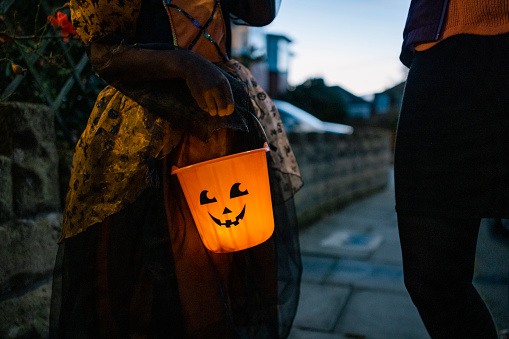 An unrecognisable girl holding a glowing orange bucket with a spooky face on it while standing on a residential street in North East England. She is ready to go trick or treating and collect sweets.