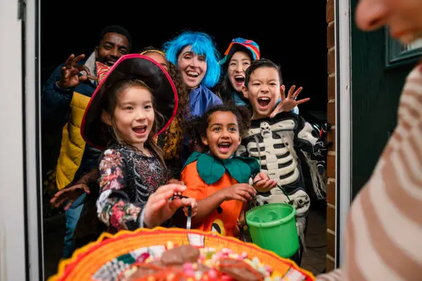 Taken from inside a residential house, two families wearing fancy dress, out trick or treating in North East England during halloween, standing outside a neighbours' house. Everyone is looking excited and are about to take sweets off a plate that an unrecognisable woman is holding.