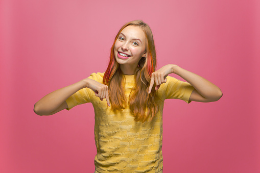 Smiling young girl pointing down recommending link, advertising, showing good sale offer on pink studio background. High quality photo