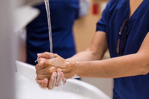 A close up photo of water streaming from a faucet as an unrecognizable healthcare worker washes her hands well with plenty of soap.
