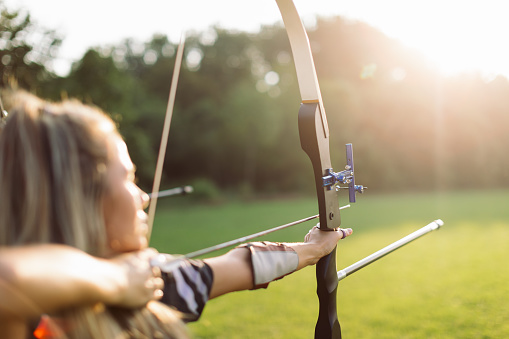 Woman practicing archery training with recurve bow on open field before sunset. She is in very good shape and focus is on the bow