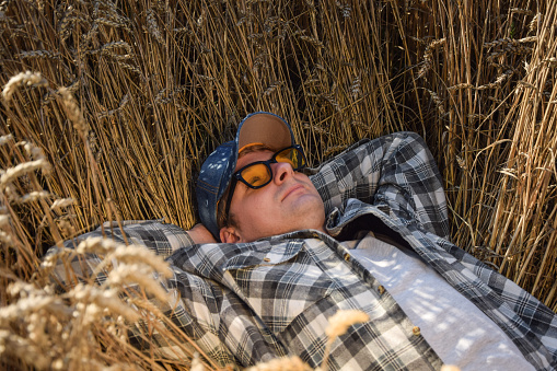Young adult farmer relaxing at agricultural grain field at sunny day. Male worker wearing hat, checkered shirt, sunglasses, lying in ripe wheat field outdoors. Countryside concept. Simple living