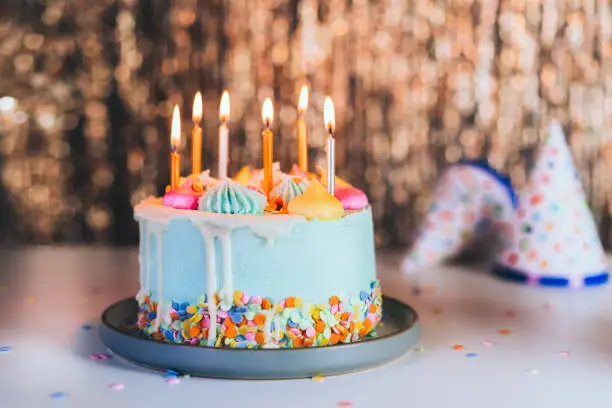 Photo of Colorful birthday cake with sprinkles and burning candles and festive caps on the sparkling gold tinsel background. Festive birthday celebration, party. Selective focus, copy space.