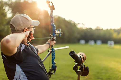 Man practicing archery training with compound bow on open field before sunset. He is in very good shape and well equipped. Focus is on his face