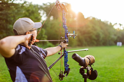 Man practicing archery training with compound bow on open field before sunset. He is in very good shape and well equipped. Focus is on his face