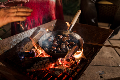 A pan full of chestnuts roasting on fire in an outdoor fire pit. There is an unrecognizable person next to the pit.