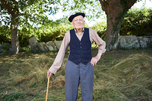 An senior grandfather poses for the camera in his casual attire with pants, vest, beret, slippers and leaning on his cane in the Galician countryside.
