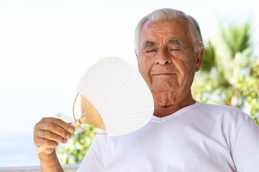 Senior man is trying to cool himself using hand fan in tropical environment. Representing heat wave solutions.