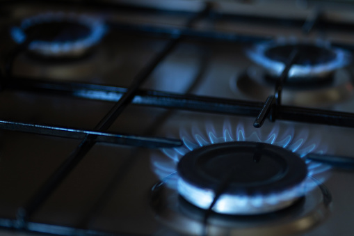 The blue flames of a gas burner on a domestic cooking hob, against a black background. The flames are reflected in the black enamel of the hob top.