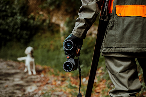 hunter looking at hunting dog with binoculars and rifle on his shoulder
