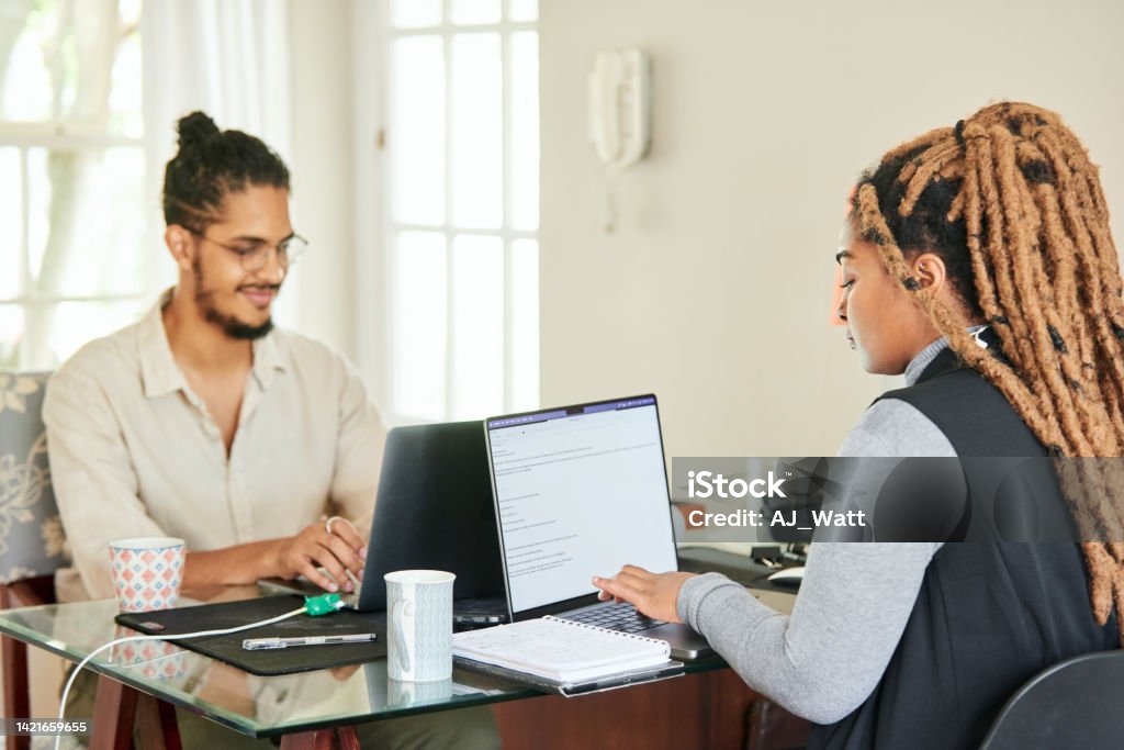 Smiling young colleagues working on laptops in their startup's home office Two smiling colleagues working on laptops while sitting at a table in their start up's home office 20-24 Years Stock Photo