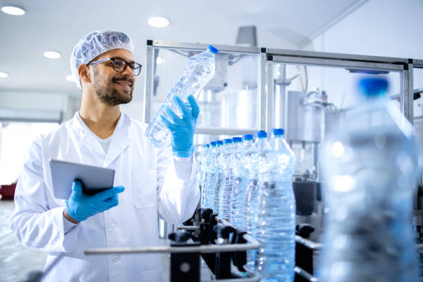 istock Technologist working in bottling factory doing quality control of drinking water before distribution. In background factory interior and industrial conveyor machine transporting PET bottles with water. 1421664420