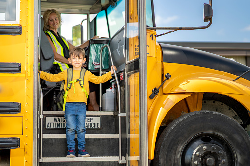 A young male Montessori student is seen stepping down off of the school bus as he arrives for the day.  He is dressed casually and has a backpack on as he looks out with a smile of anticipation.