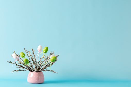 Easter eggs decorations and pussy willow twigs with catkins in a vase. Blue background, space for copy.