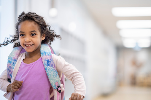 A sweet Montessori student runs through the hall of her school as she makes her way out at the end of the day.  She is dressed casually and has a backpack on as she smiles and makes her way out.