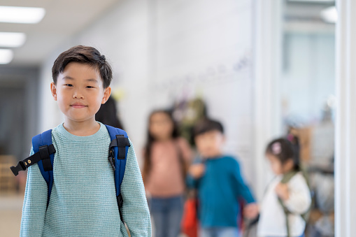 A young Montessori student of Asian decent is seen standing in the hallway as he poses for a portrait.  He is dressed casually and has a backpack on and his peers can be seen in the background.