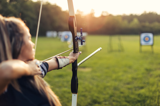 Woman practicing archery training with recurve bow on open field before sunset. She is in very good shape and focus is on the bow