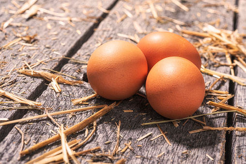 Three farm fresh brown chicken eggs in an old rusty wire basket sitting on old weathered wooden planks, with pieces of hay scattered around. The bottom of the basket is lined with a layer of hay to protect the eggs.