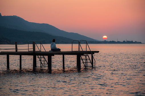 Silhouette of a woman practicing yoga alone on a pier at sunrise. Yoga, fitness, healing and a healthy lifestyle.