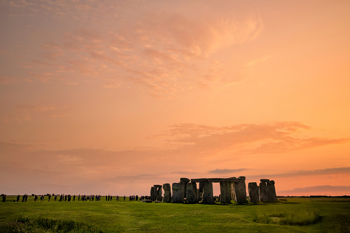 Stonehenge during a bright orange sunset with a line of people in the distance.