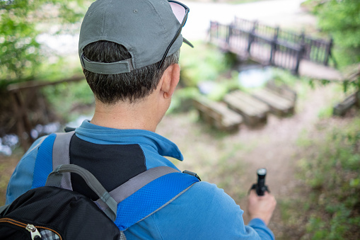 A middle-aged man is engaged in mountain tourism. He is wearing a blue tracksuit with a blue, gray, and black backpack, a gray baseball cap, gray trainers, and a walking stick in his hand. A mountain hiker descends a steep trail to a small wooden bridge in the mountain. Back view.