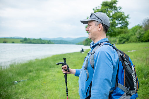 A middle-aged man is engaged in mountain tourism. He is wearing a blue tracksuit with a blue, gray and black backpack, a gray baseball cap, gray trainers and a walking stick in his hand. Mountain hiker by a lake in the mountains.