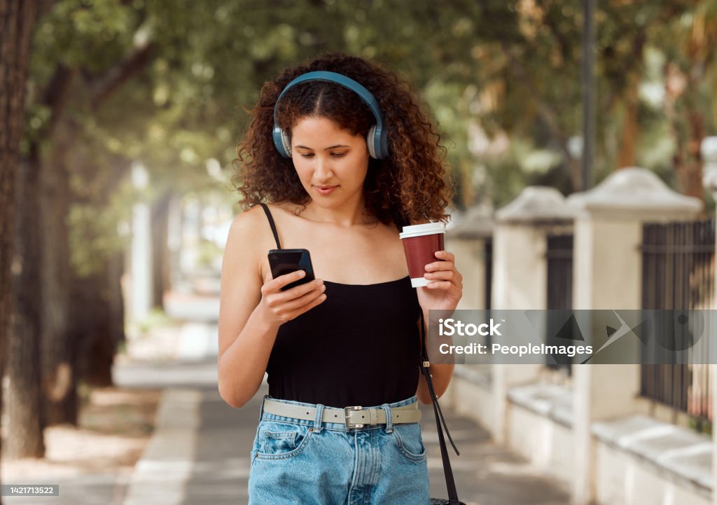 One young mixed race woman standing in the city and using her cellphone to listen to music through headphones while drinking a takeaway coffee. Hispanic woman browsing the internet on a phone downtown Music Streaming Service Stock Photo