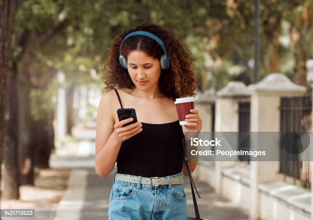 One Young Mixed Race Woman Standing In The City And Using Her Cellphone To Listen To Music Through Headphones While Drinking A Takeaway Coffee Hispanic Woman Browsing The Internet On A Phone Downtown Stock Photo - Download Image Now