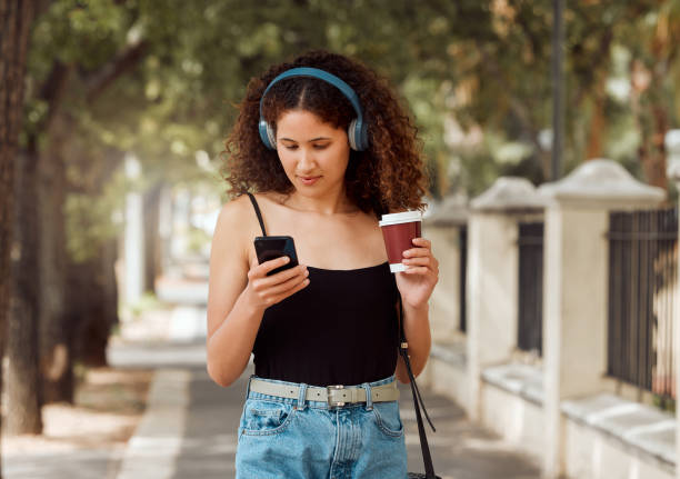 pewna młoda kobieta mieszanej rasy stojąca w mieście i używająca telefonu komórkowego do słuchania muzyki przez słuchawki podczas picia kawy na wynos. latynoska kobieta przeglądająca internet na telefonie w centrum miasta - spotify zdjęcia i obrazy z banku zdjęć