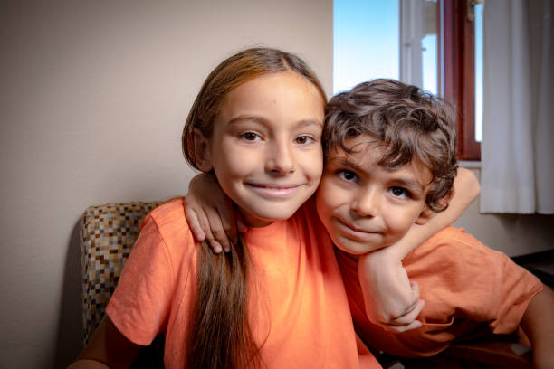 11 year old girl with straight long hair. Smiling face.6 years old boy. Big brother and sister hug each other. Moments full of love. Inside the house.