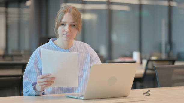 istock Young Woman with Laptop Reading Documents in Office 1421725878