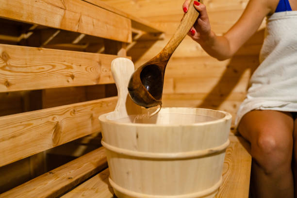 hand of unknown caucasian woman in sauna spa taking water from bucket with wooden spoon to pour on the hot stones stock photo