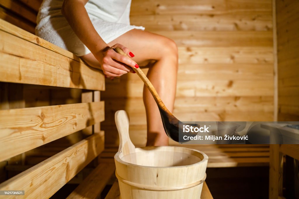 hand of unknown caucasian woman in sauna spa taking water from bucket with wooden spoon to pour on the hot stones Sauna Stock Photo