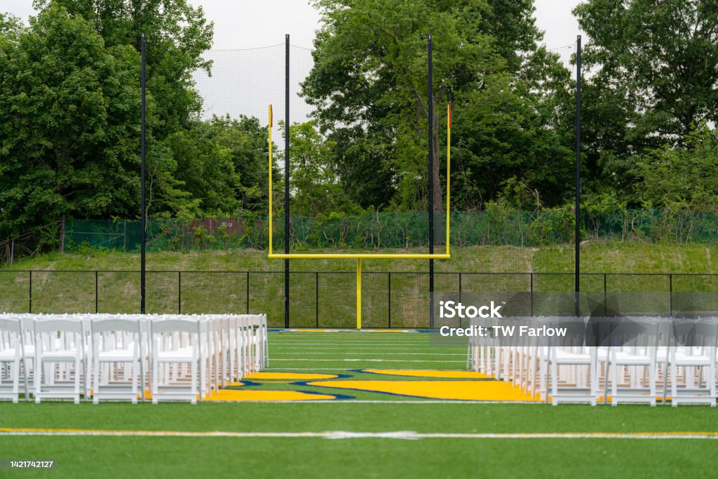 White chairs set-up in rows on a green synthetic turf athletic field for a high school graduation ceremony. Artificial Stock Photo