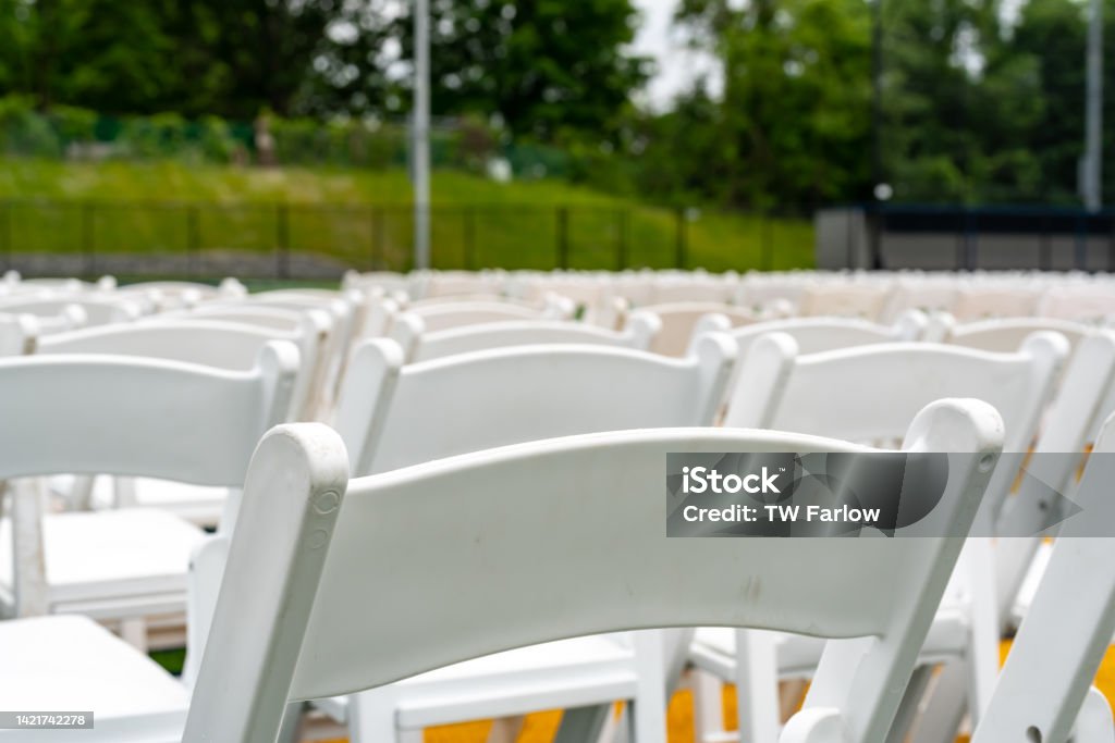 White chairs set-up in rows on a green synthetic turf athletic field for a high school graduation ceremony. Artificial Stock Photo