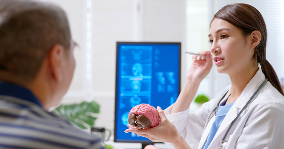 attractive young brunette ponytail female asian doctor wear white coat and stethoscope pen pointing explaining brain model to gray hair old sick at clinic - an xray film display on computer