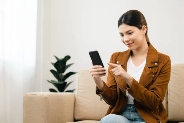 istock Beautiful young women using mobile phone sitting on sofa in living room. Using smartphone. Woman Shopping online store. Relax time at home 1421755169