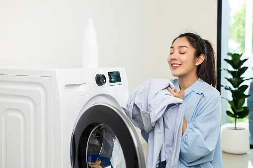 Happy housewife opens the front of the washing machine and smiles to receive the laundry that has been washed at house. Woman smelling fragrant clean clothes washed from softener and washing machine.