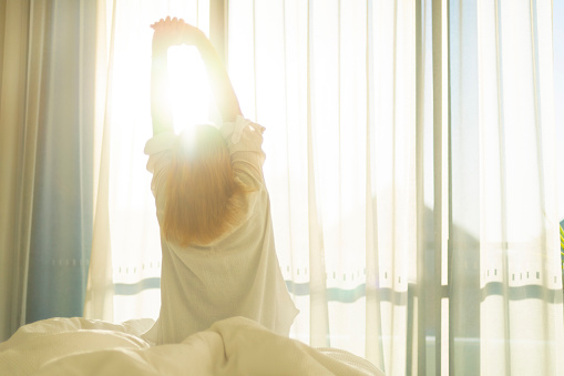 Woman wearing nightwear waking and stretching in the morning light. There is a curtained window behind her with sunlight streaming in