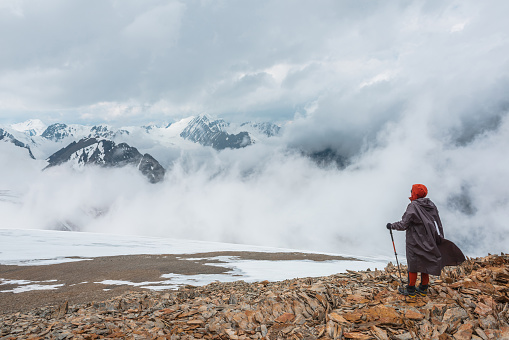 Hiker and high snowy mountains in dense low clouds. Minimal scenery with man on stone hill against beautiful mountain peaks in thick clouds. Simple minimalism with man and majestic mountains nature.