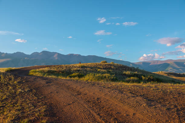 Tourist on sunlit gold grassy hill with dirt road with view to high mountain range. Beautiful sunny mountain landscape with man in mountains. Colorful scenery with traveler on hill in golden sunlight. Tourist on sunlit gold grassy hill with dirt road with view to high mountain range. Beautiful sunny mountain landscape with man in mountains. Colorful scenery with traveler on hill in golden sunlight. western siberia stock pictures, royalty-free photos & images