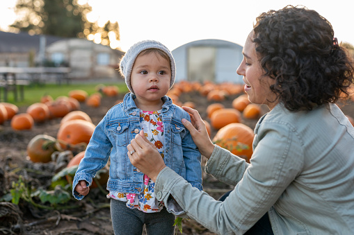 A fit ethnic mother in her thirties is buttoning the jean jacket on her 18 month old daughter at a pumpkin patch. The girl appears to be happy and relaxed.