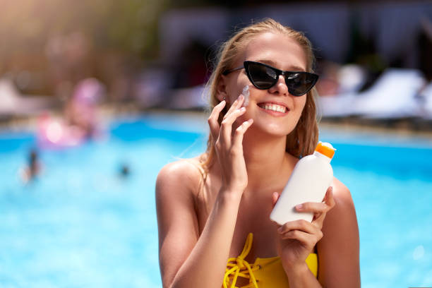 Blonde woman applies sunscreen solar cream on cheeck and face near swimming pool. Smiling pretty girl puts suntan cream from plastic bottle on skin in spa. Female in bikini with suntan lotion. stock photo